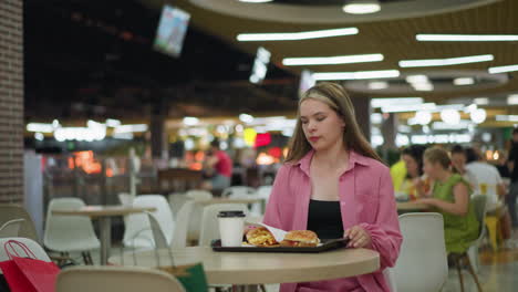 lady in pink dress sits down, prepares to eat her breakfast with a serious expression on her face, tray with coffee, burger, and fries on the table, blurred background with bokeh lighting