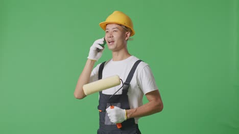 Side View Of Asian Man Painter Wearing Safety Helmet Talking On Smartphone And Looking Around While Standing In The Green Screen Background Studio