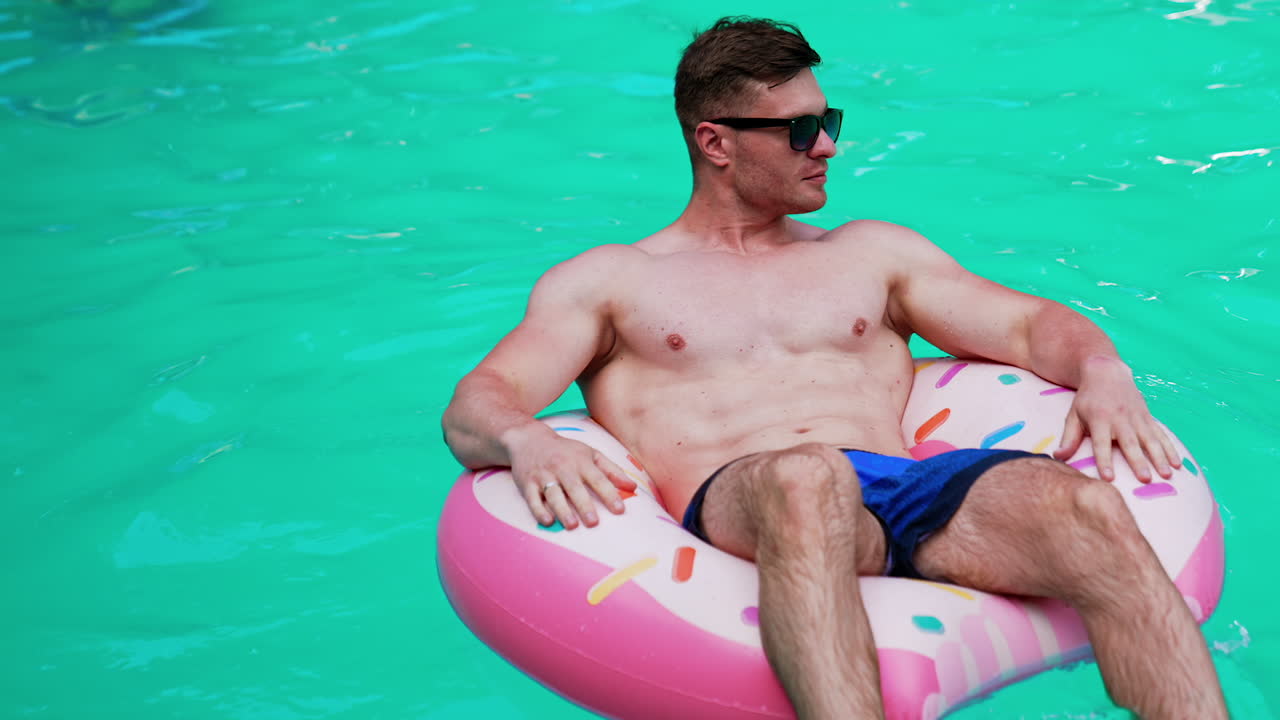 Vacation time in the city. Handsome Caucasian athletic man wearing sunglasses swims sitting in the inflatable donut.