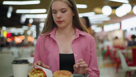 woman in pink dress sits down in a restaurant, fries, burger, and coffee in a tray, she reaches for fries with bokeh lighting and a blurred background filled with people in the mall