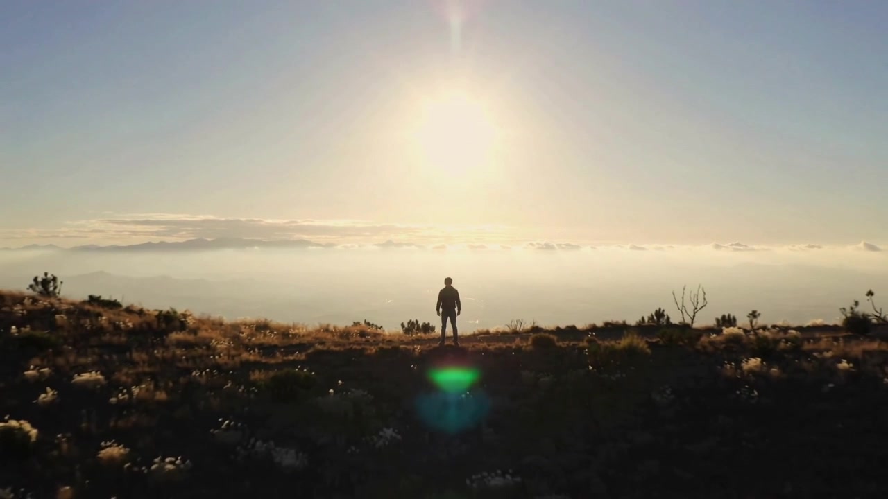 Sunrise drawing jacketed hiker standing on mountain ridge, watching valley with green lens flare