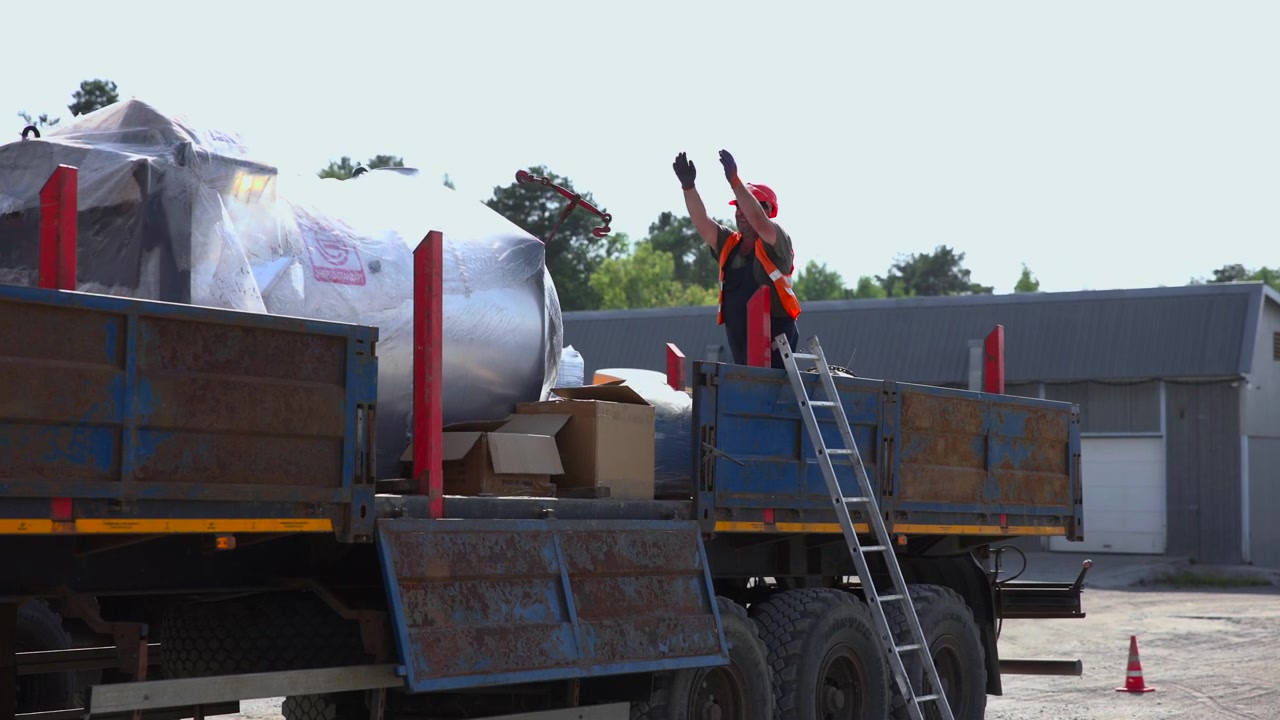 Worker in red hard hat and orange safety vest stands on flatbed truck beside metal ladder, with covered cargo, cardboard boxes, and weathered metal sides in bright industrial yard, warehouse