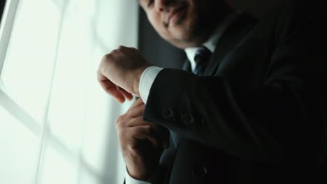 confident businessman in classic blue suit buttoning or adjust cufflinks near window in hotel room at the morning. handsome man wearing a nice suit on wedding day.