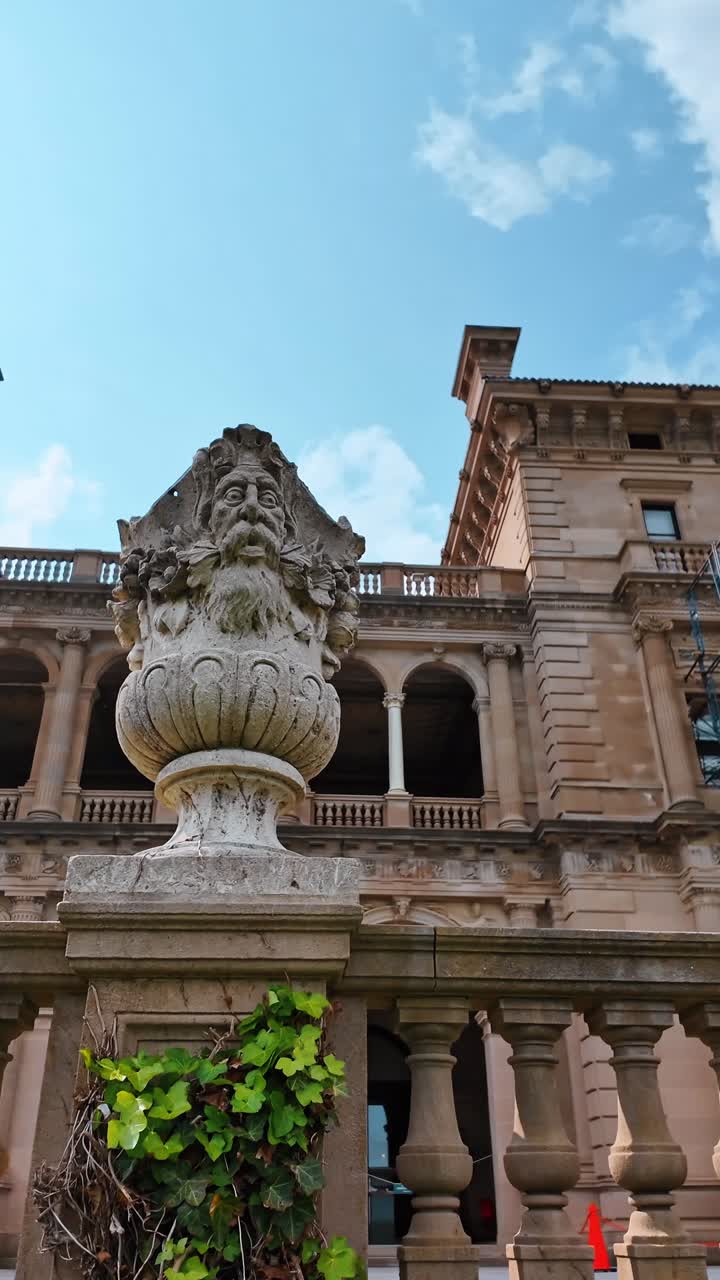 Scultura architettonica in pietra su piedistallo con sfondo di palazzo. Urna in pietra con dettaglio di viso intagliato posizionata su una balaustra con un grande edificio storico dietro.