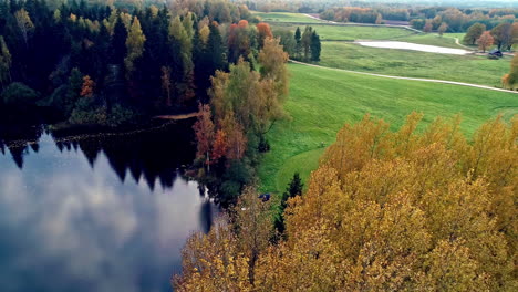 Scenic-aerial-shot-over-rectangular-cabin-beside-a-pristine-lake-surrounded-by-autumnal-trees-in-rural-countryside-at-daytime