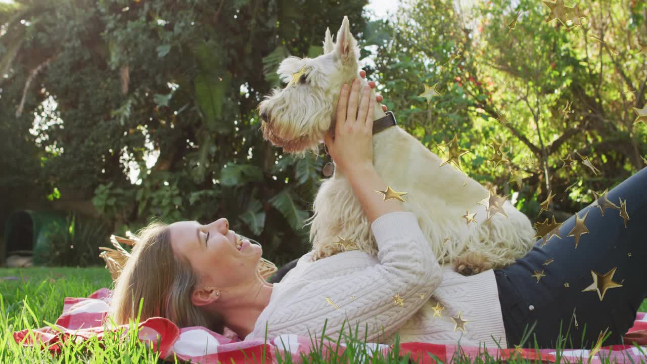 Woman lifting terrier onto torso after lick and stroking ears on picnic blanket for pet care