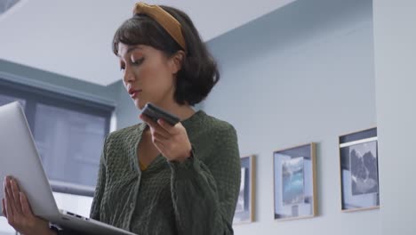 Biracial-businesswoman-talking-on-smartphone-and-using-laptop-alone-at-office