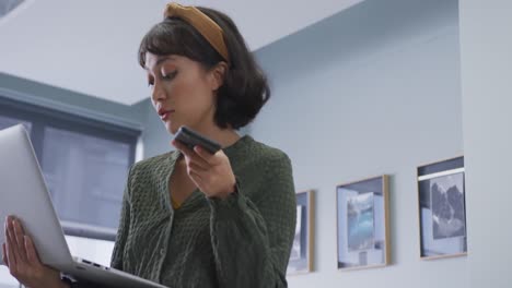 mujer de negocios biracial hablando en teléfono inteligente y usando portátil sola en la oficina