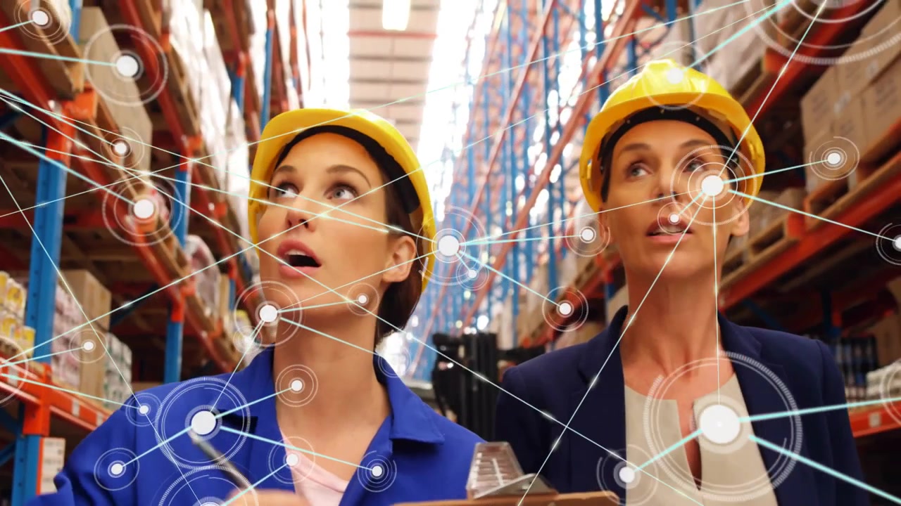 Giovane donna con casco che punta una penna verso le scatole richiamando una sovrapposizione dell'inventario, mentre un collega scansiona gli scaffali