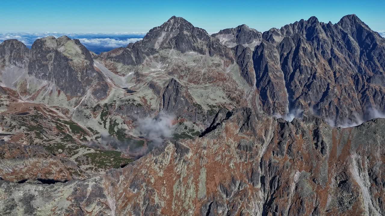 Hyperlapse drone shot flying over a rugged ridge in the High Tatras, gradually exposing a deep cloudy valley surrounded by towering peaks under clear skies