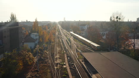 Scenic-Aerial-Shot-of-Passenger-Train-driving-into-Sunset-on-Train-Tracks-surrounded-by-Trees-above-Cityscape-in-Berlin-Germany,-Dolly-forward