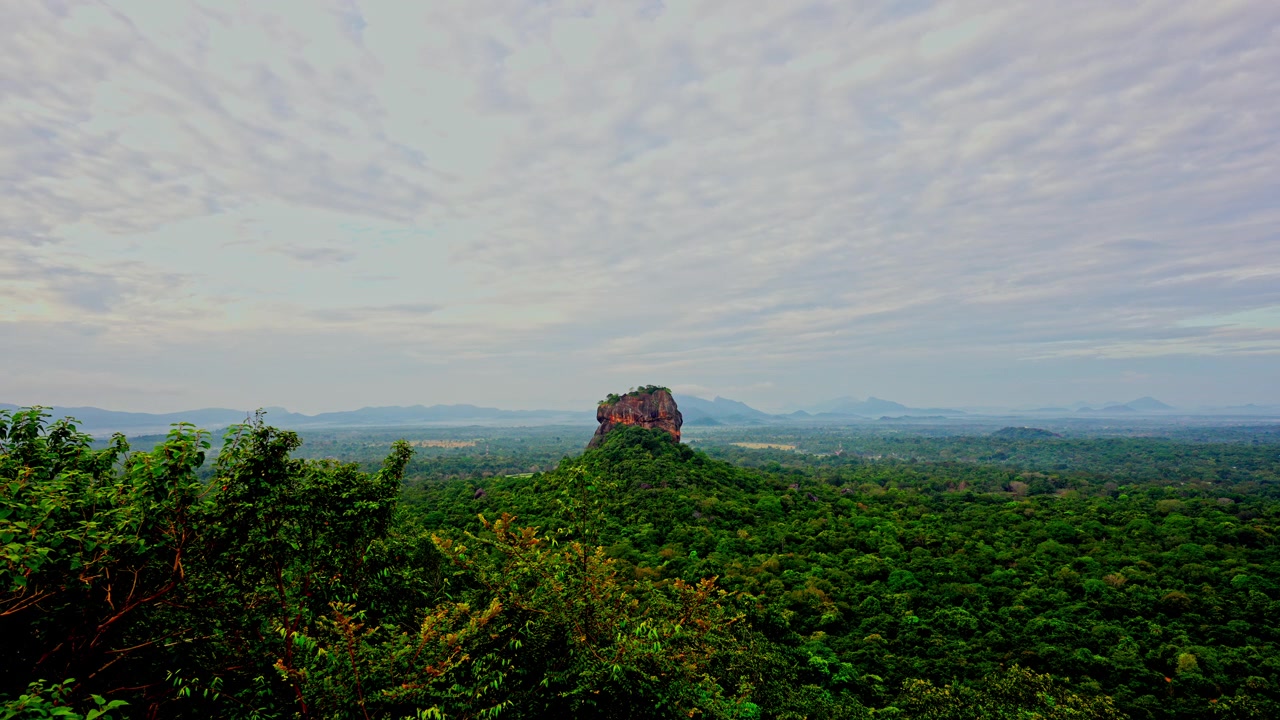Ampia vista della fortezza rocciosa di Sigiriya che emerge dalla giungla nebbiosa all'alba, con nuvole leggere che solcano il cielo sopra le pianure dello Sri Lanka centrale.