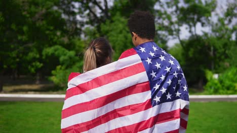 Vista-Posterior-De-Una-Atractiva-Pareja-Multiétnica-Abrazándose-Sosteniendo-La-Bandera-Americana-En-La-Espalda-Y-Caminando-En-El-Campo-Verde