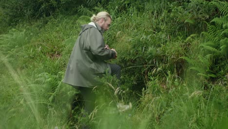 young, long haired, bearded, caucasian male filling waterbottle from natural stream water source and drinking in lush green scandinavian nature