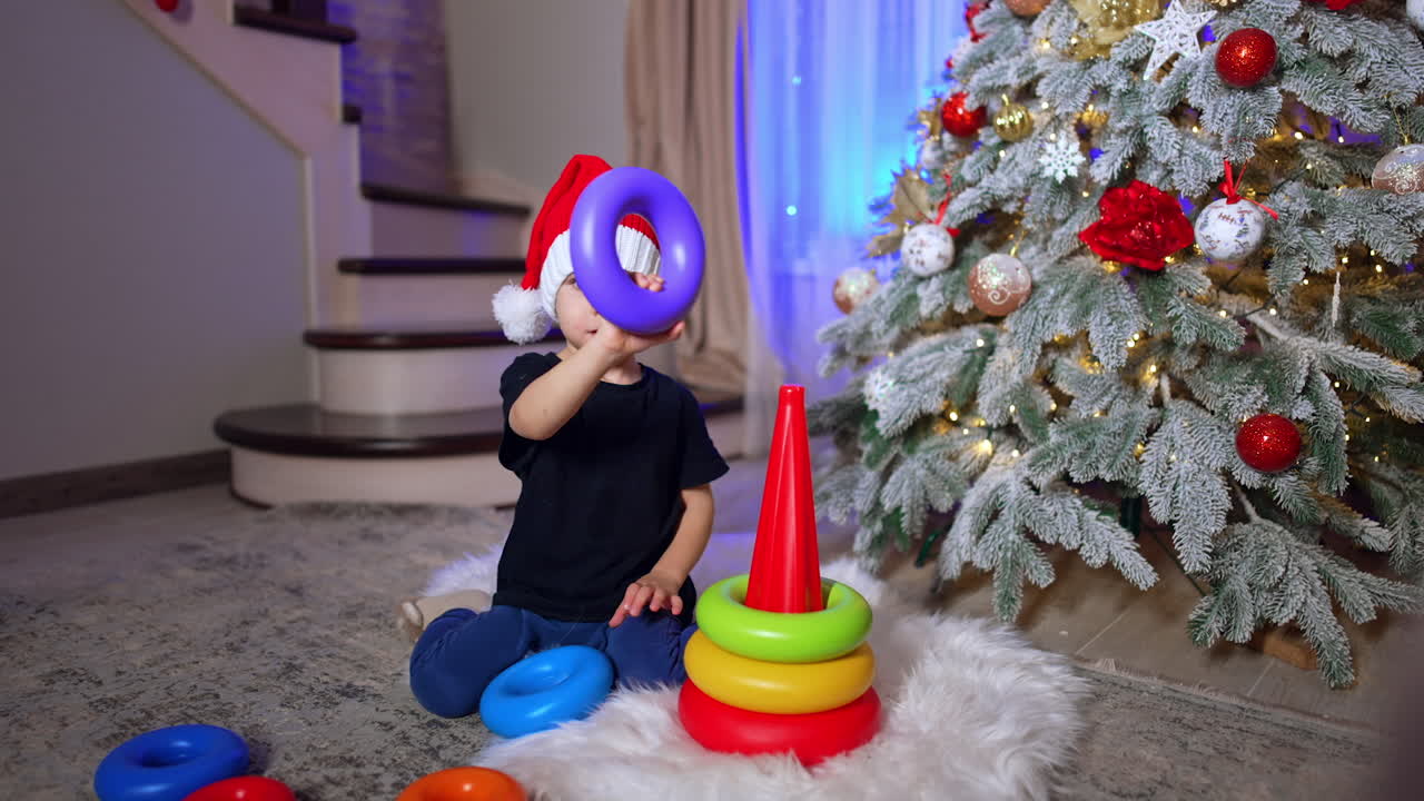 Two year old toddler in Santa cap plays with pyramid. Lovely child having fun at home at Christmas.