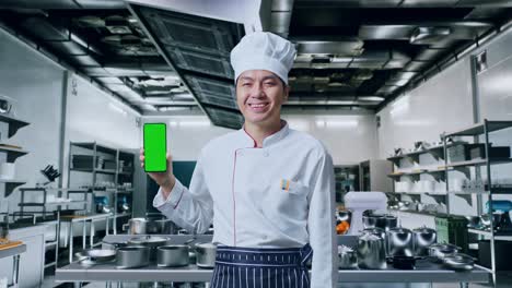 un chef asiático sonriendo y mostrando un teléfono inteligente de pantalla verde a la cámara mientras está de pie en la cocina