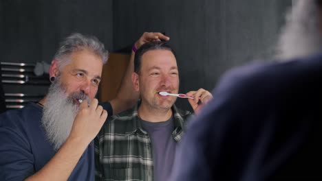 Happy-LGBT-couple-an-elderly-man-with-gray-hair-and-a-lush-beard-together-with-his-brunette-boyfriend-with-stubble-in-a-green-checkered-shirt-brush-their-teeth-with-a-toothbrush-in-the-bathroom-of-a-modern-apartment-in-the-morning