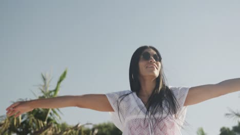 Feliz-Mujer-Hispana-Con-Gafas-De-Sol-Bailando-En-La-Playa-Soleada,-Sonriendo,-Cámara-Lenta