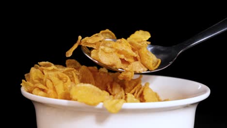 a spoon lifts cornflakes from a white bowl against a black background, highlighting the cereal's texture and movement