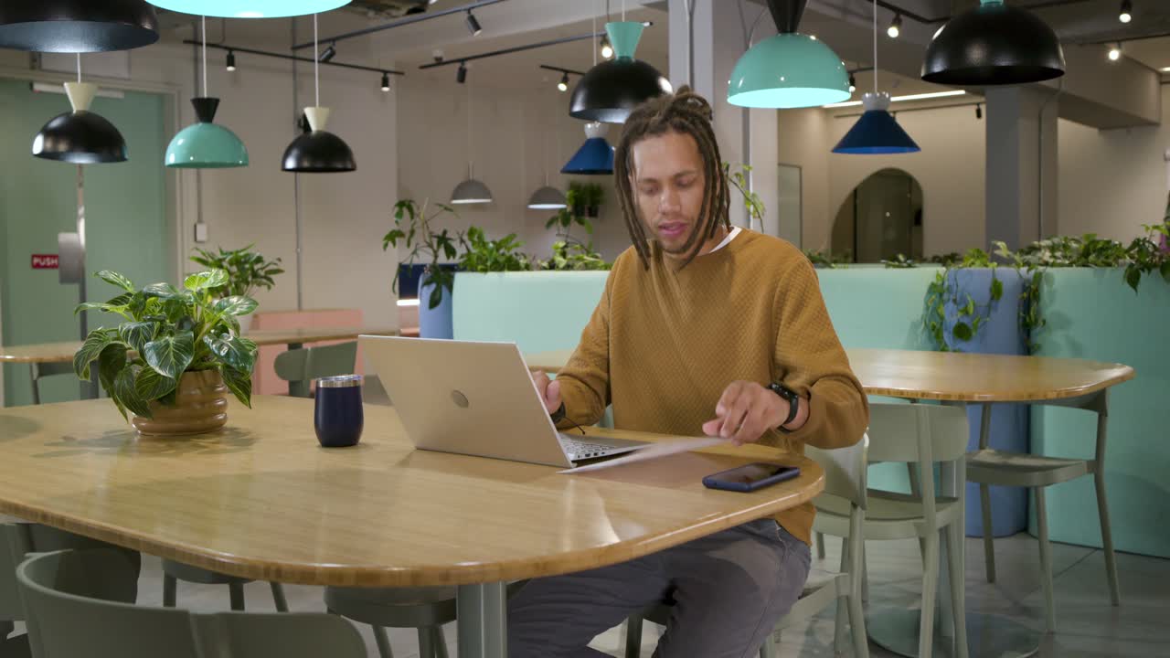 Man in sweater finding confusing pages, calling phone while typing laptop at shared workspace table