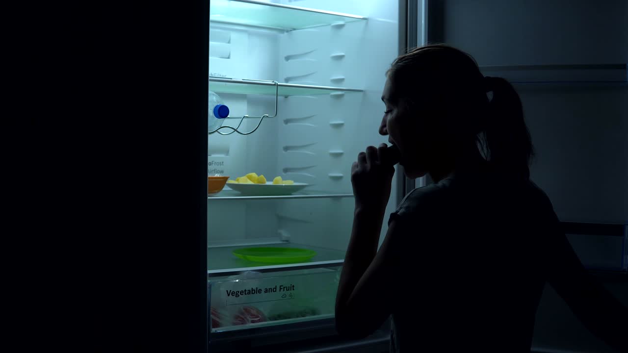 Di spalle, una giovane ragazza affamata prende un cetriolo dal frigorifero per mangiarlo di notte.