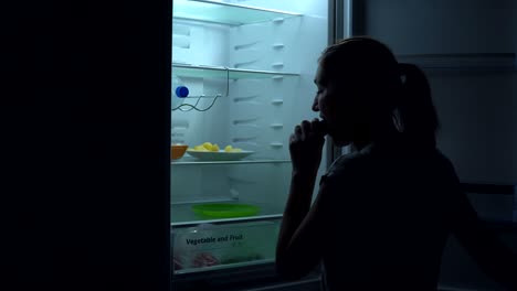 rear view of young girl is hungry and takes cucumber from the refrigerator to have bite at night