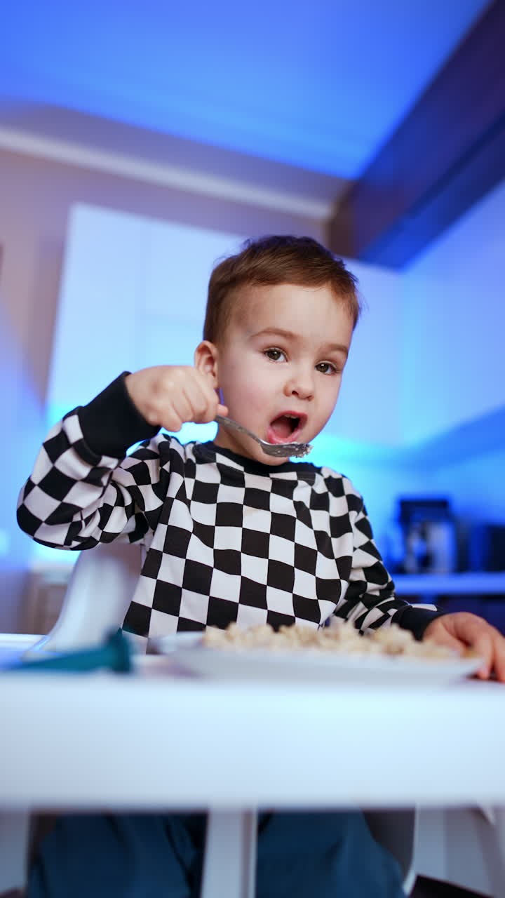 Sweet little kid in checkered shirt picking some porridge with a spoon. Low angle view on the baby eating himself at home. Vertical video.