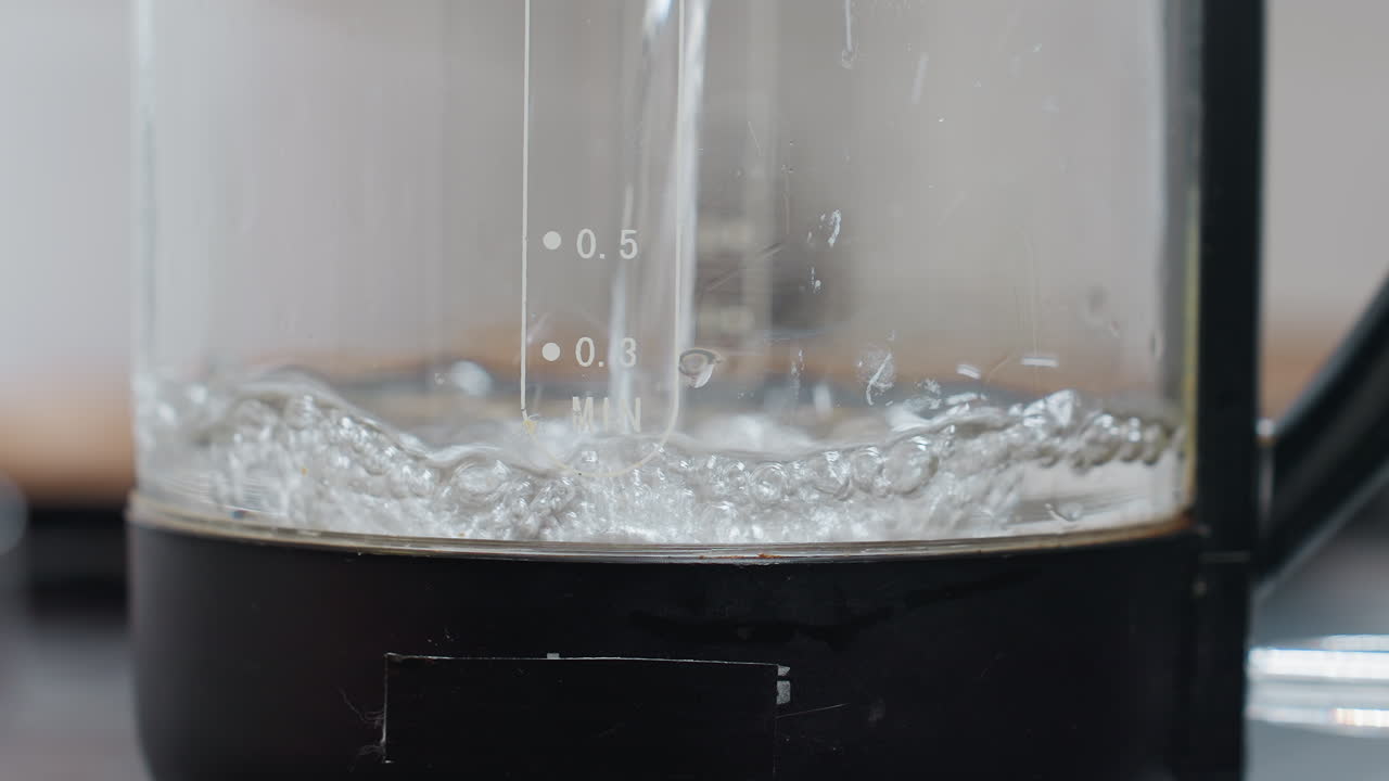 電気ジャーに水を入れているクローズアップ。ジャーには文字が刻まれており、液体で満たされ、透明な水の泡ができている様子が、モダンなキッチンを背景に映し出されています。