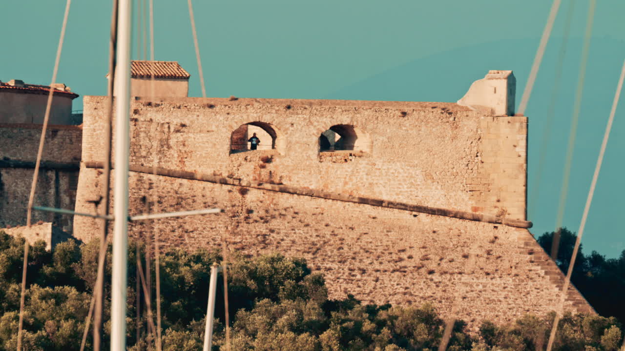 A close up of an ancient stone fortress wall with arched windows, framed by tall sailboat masts