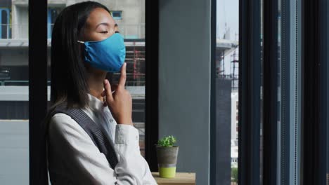 mujer asiática pensativa con máscara facial mirando por la ventana en una oficina moderna
