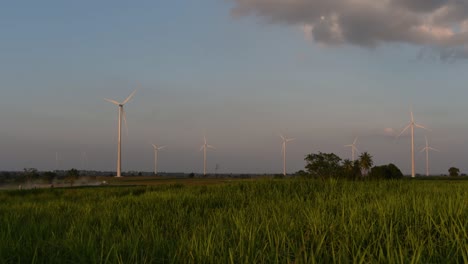 Wind-Turbines-shooting-out-of-a-Farmland-while-the-Sun-is-setting-as-Trucks-Pass-by-making-Clouds-of-Dust,-clean-alternative-energy-in-Thailand-and-mainland-Southeast-Asia