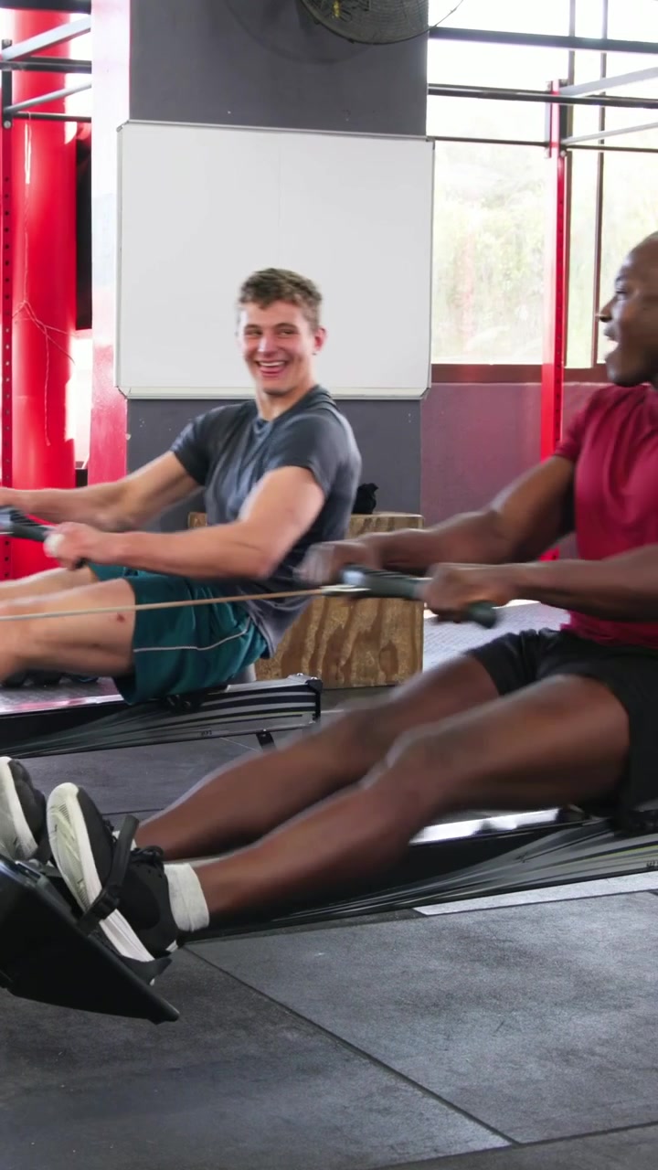 Vertical video: Rowing two men red shirt leading, starting at catch pulling on ergs at gym training