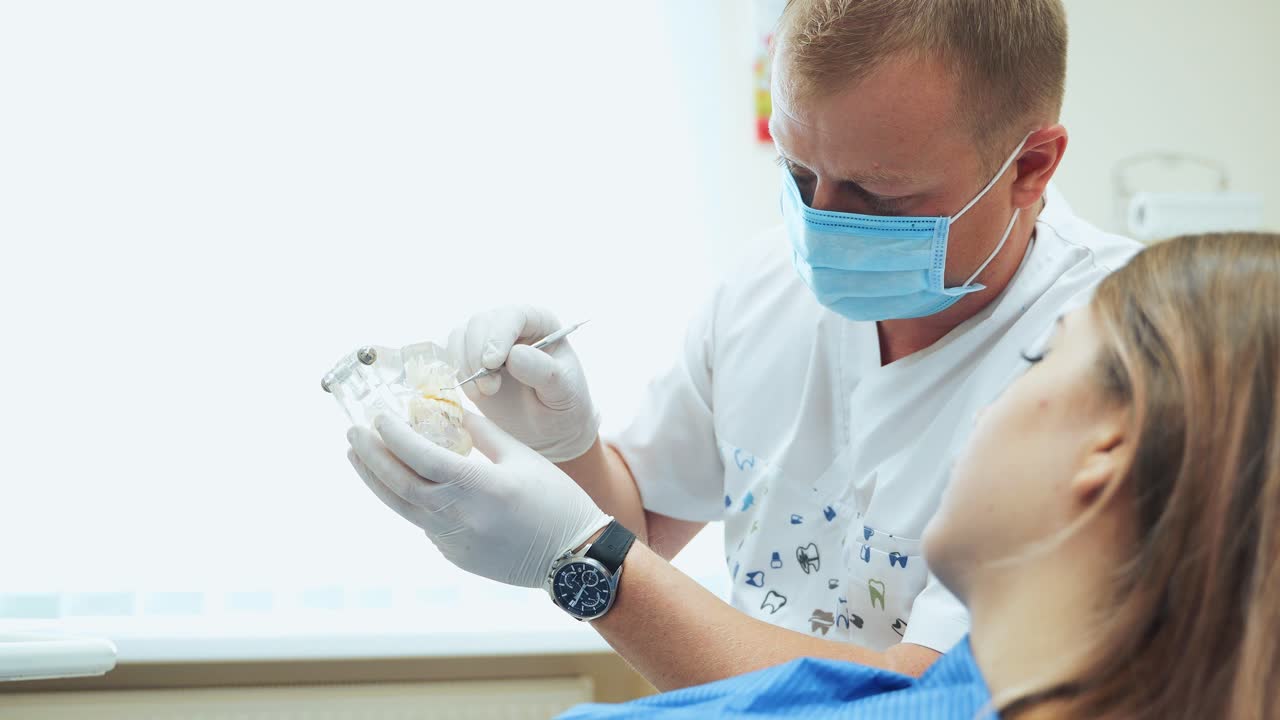 A male dentist shows a girl a jaw layout. Visit to the doctor in a mask and white gloves
