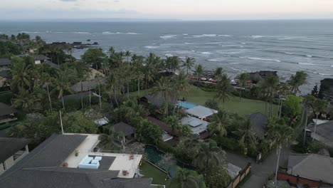 Low-aerial-skims-over-homes-and-resorts-to-volcanic-beach,-Cemagi-Bali
