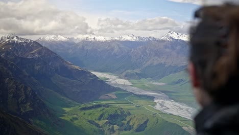 Girl-looking-over-a-beautiful-green-valley-with-mountains-with-a-river-running-through-on-a-bright-sunny-day-on-a-grassy-hill,-drifting-handheld-closeup-shot