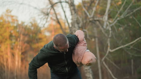 a joyful moment captured in a sunny outdoor park where a man in a green jacket playfully spins a little girl in a pink jacket and blue jeans, both smiling and enjoying the cheerful atmosphere