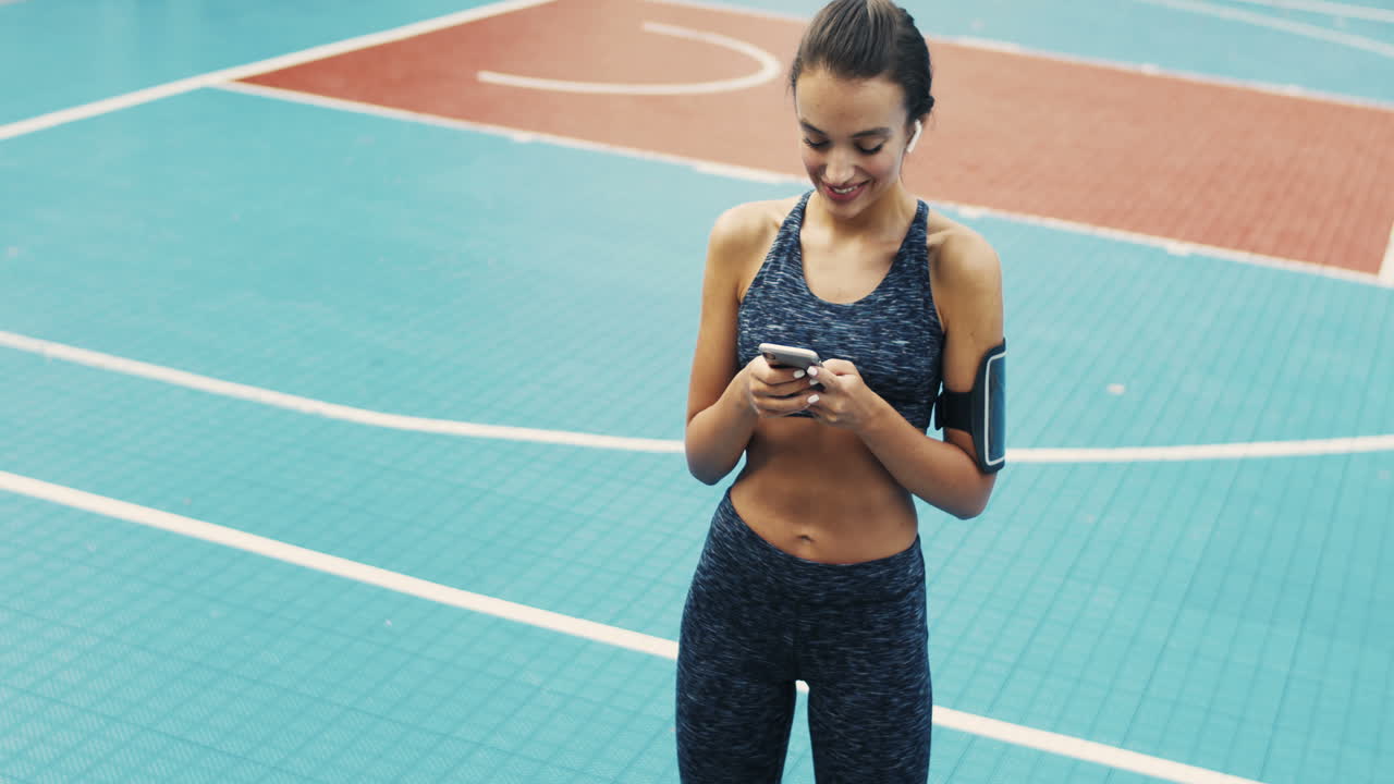 chica deportiva con mensajes de texto de airpods en el teléfono inteligente mientras está de pie en la cancha al aire libre en un día de verano