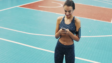 Sportliches-Mädchen-Mit-Airpods-SMS-Nachricht-Auf-Dem-Smartphone,-Während-Es-An-Einem-Sommertag-Auf-Dem-Außenplatz-Steht