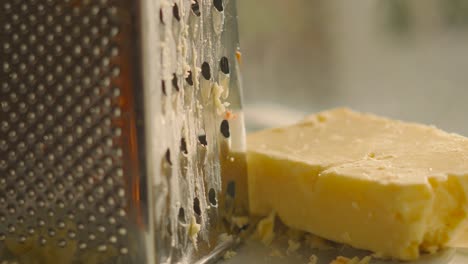 descending camera view of grater after grating cheddar with block of rustic cheese resting on plate in natural light. dairy based food product