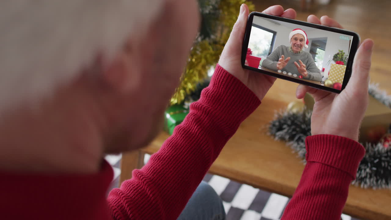 hombre caucásico con sombrero de santa usando un teléfono inteligente para una videollamada de navidad con un hombre sonriente en la pantalla