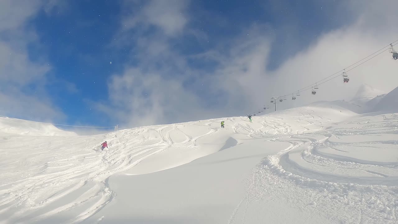 Group of skiers descending powder off-piste, leaving tracks, with chairlift in background on clear sunny day