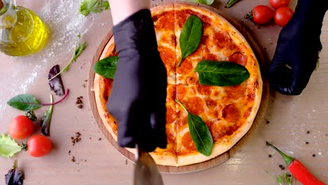 cook cuts pizza salami with spinach leaves on slices, hands in rubber gloves.