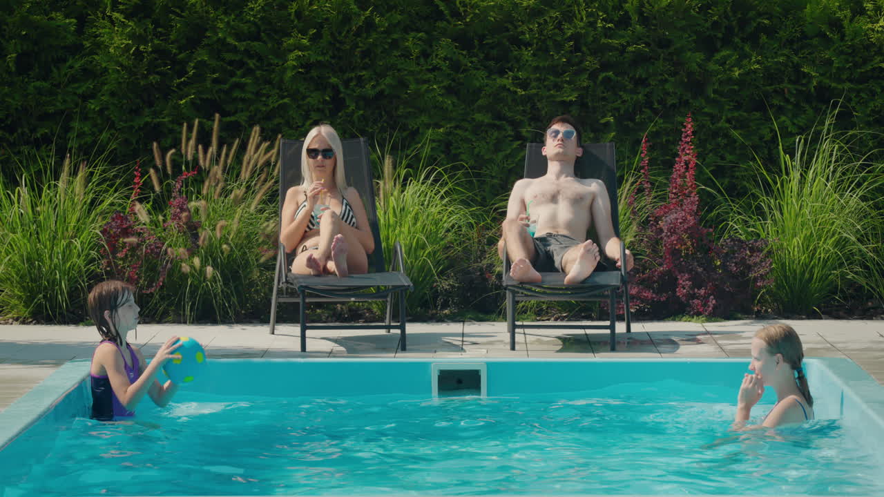 The family relaxes by their pool in the backyard of the house. Adults sunbathe on sun loungers, two children swim in the pool