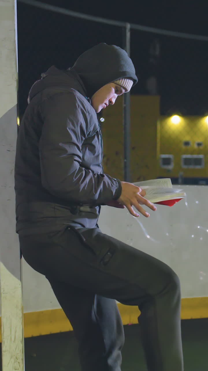 atleta con capucha apoyado contra un poste de meta metálico al aire libre por la noche volteando páginas de un libro con una pelota de fútbol rodando suavemente bajo los pies, iluminado por las cálidas luces de la ciudad urbana