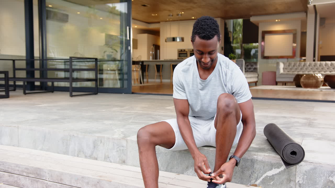 Hombre afroamericano sentado en los escalones, con ropa deportiva, relajándose con una esterilla de yoga cerca, en casa.