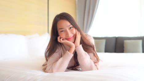 portrait of young smiling asian woman lying on bed, leaning head on hands and looking at camera, slow motion