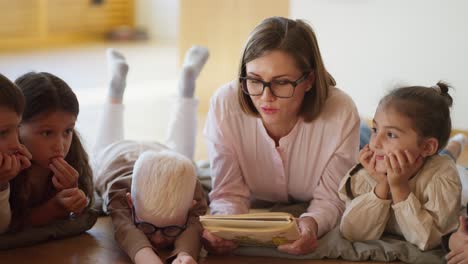 Close-up-shot-of-a-blonde-woman-with-glasses-in-a-pink-shirt-reading-a-book-to-children-in-a-club-for-preparing-children-for-school-in-a-special-room-Lying-on-pillows-on-the-floor