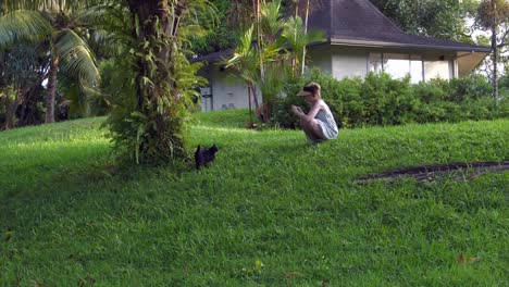 Gato-Corriendo-Por-Una-Colina-Cubierta-De-Hierba-Hacia-Una-Hembra-Blanca-En-Cuclillas-Y-Filmando-Y-Luego-Acariciando-Al-Gato