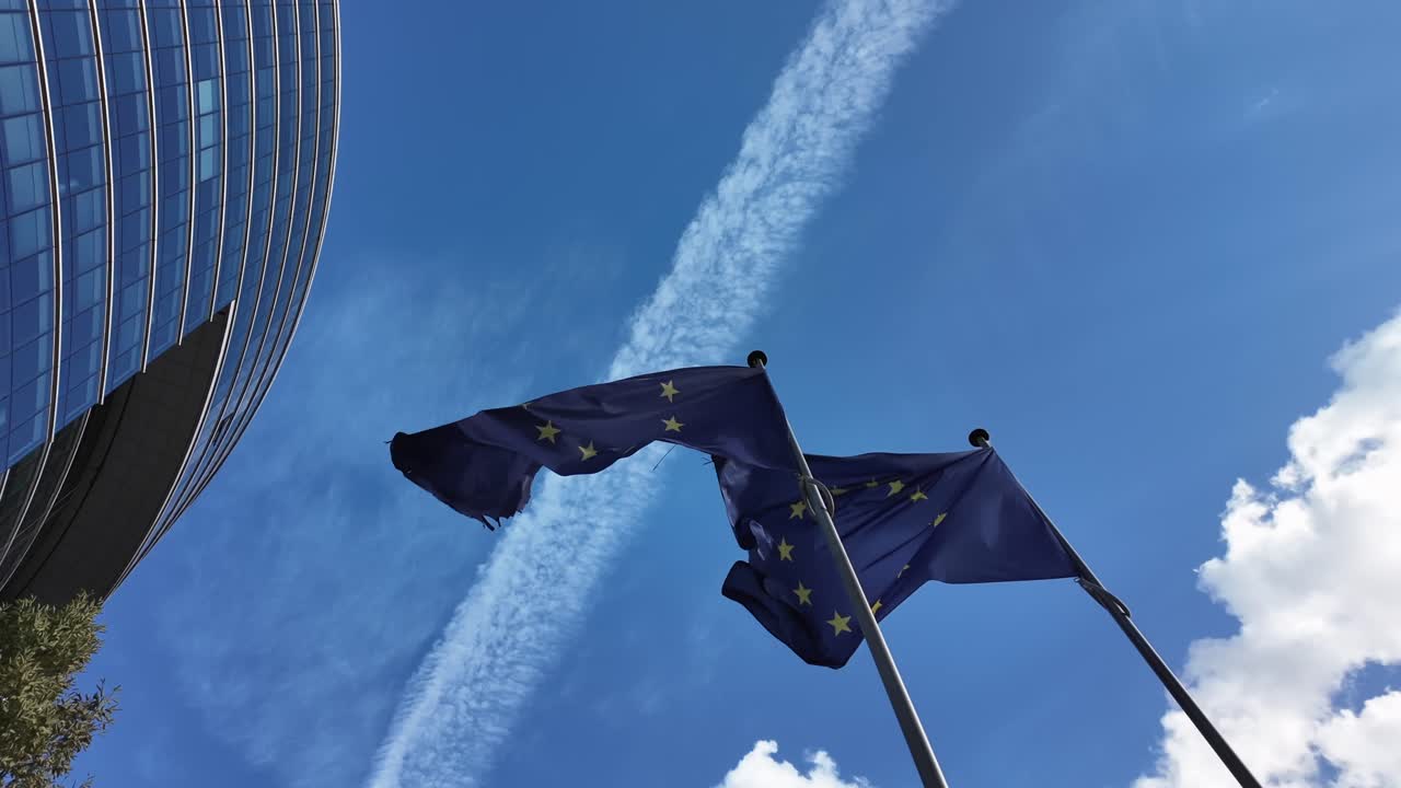 European Union flags flutter under a clear sky with airplane contrail and the Lex Building in Brussels’ European Quarter
