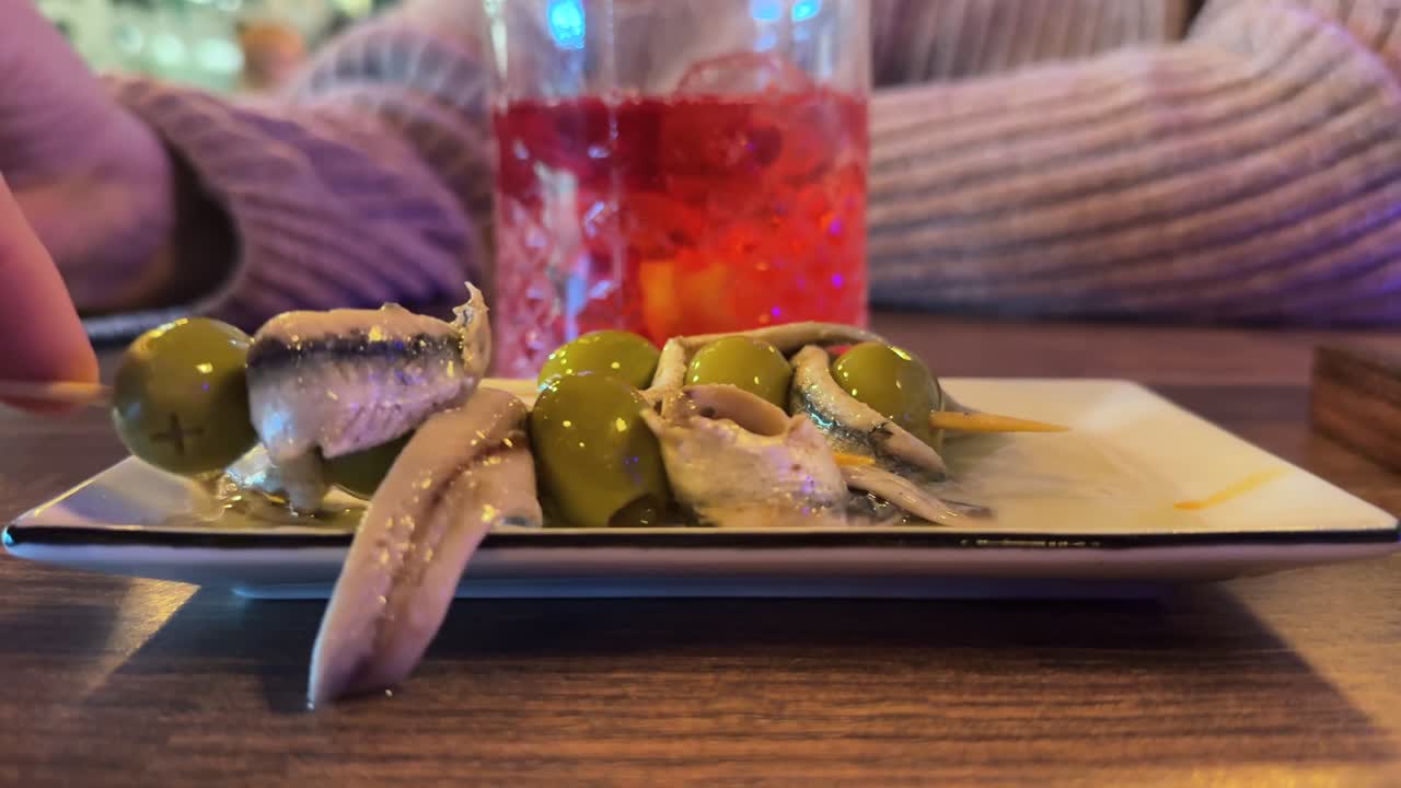 Primissimo piano della mano di un uomo che prende uno spiedino di olive verdi e acciughe all'interno di un tradizionale bar spagnolo
